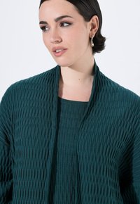 Tyrkysový, strukturovaný pletený cardigan s ribbed vzorem, otevřeným předním dílem a uvolněným střihem. Má kulatý výstřih a jemně splývá.