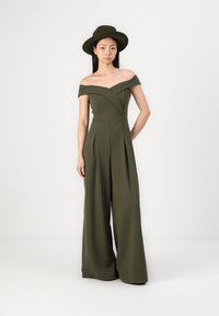 Salopette verde oliva senza spalline con gambe larghissime plissettate. Abbinata a un cappello in feltro aderente, che mette in risalto il tessuto liscio e le linee pulite.