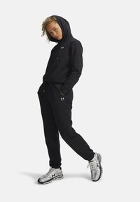 Felpa nera con cappuccio e coulisse regolabili, abbinata a pantaloni jogger neri. Presenta un marchio argentato e un tessuto confortevole. Scarpe sportive argentate.