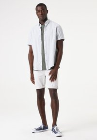 Camisa de manga corta gris claro, camiseta verde debajo, pantalones cortos blancos y zapatillas azules con suelas blancas. Tela suave y diseño casual.