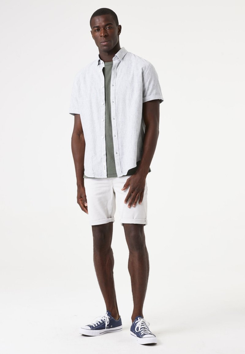 Camisa de manga corta gris claro, camiseta verde debajo, pantalones cortos blancos y zapatillas azules con suelas blancas. Tela suave y diseño casual.