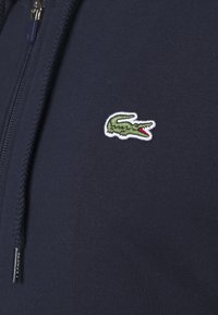 Tmavě modrá zipová mikina vyrobená z bavlny. Obsahuje malé, vyšívané zelené logo krokodýla na hrudi. Texturovaná tkanina, nastavitelná kapuce.