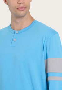 Camicia a maniche lunghe di colore blu chiaro con colletto henley e due bottoni. Presenta accenti a strisce grigie sulle maniche. Tessuto morbido e vestibilità rilassata.