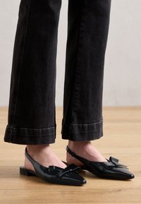 Bailarinas negras tipo slingback con puntera afilada, con un acabado brillante y un lazo decorativo en la parte superior, combinadas con jeans oscuros de corte recortado.