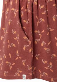 Jupe en tissu bordeaux doux avec un petit motif floral de feuilles et baies orange et une poche latérale avec une étiquette de marque blanche à l'ourlet.
