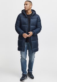 Cappotto invernale blu navy con cappuccio, caratterizzato da un design trapuntato, zip frontale e polsini elasticizzati, abbinato a jeans blu e scarpe sportive scure.