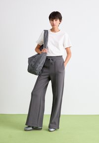 Jeune personne portant un t-shirt blanc, un pantalon de jogging large gris, des sneakers argentées, et portant une grande sacoche grise sur un fond de sol vert.