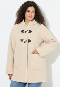 Béžový kabát s kapucí, s texturovaným povrchem, dvěma předními kapsami a zapínáním na šňůrky. Dlouhé rukávy s uvolněným střihem, v kombinaci s džínami.