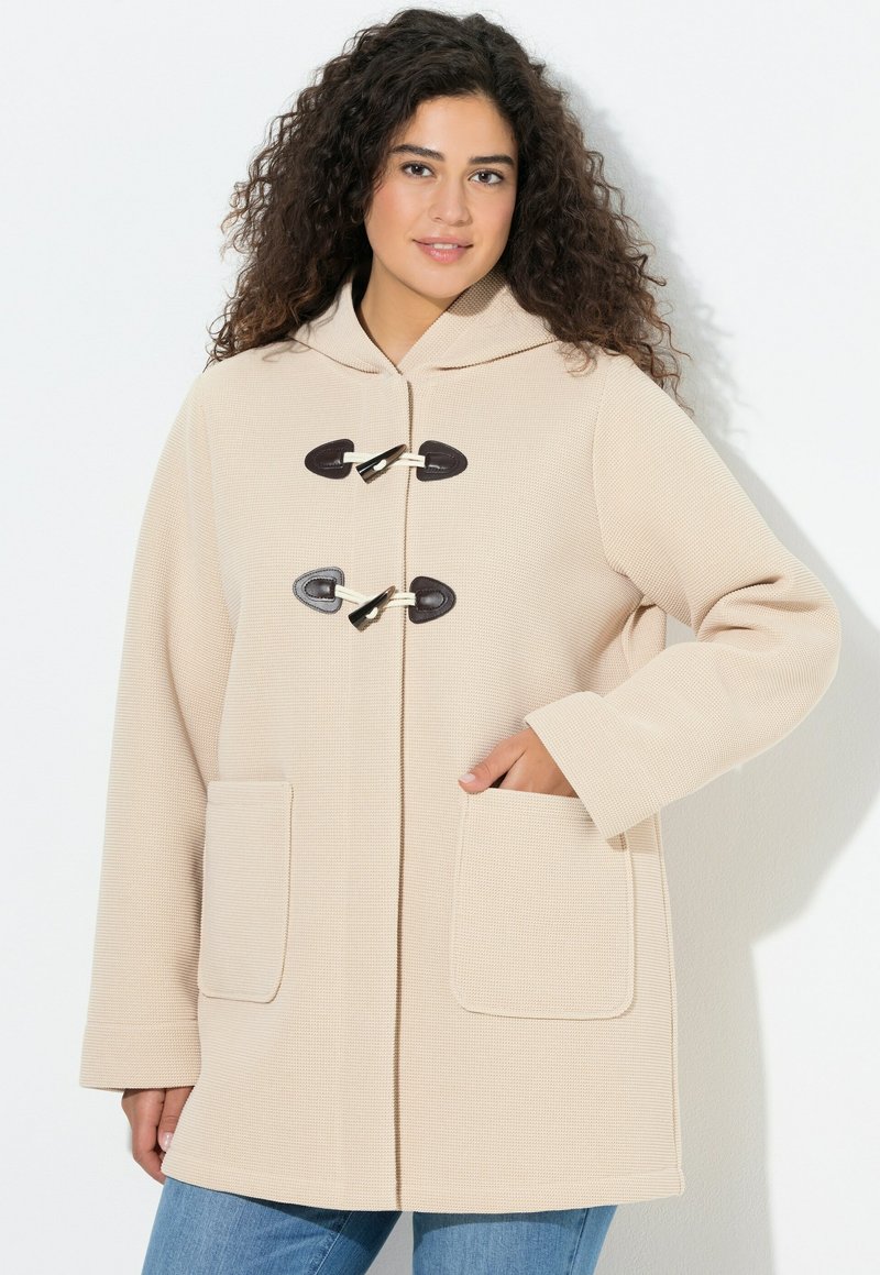 Béžový kabát s kapucí, s texturovaným povrchem, dvěma předními kapsami a zapínáním na šňůrky. Dlouhé rukávy s uvolněným střihem, v kombinaci s džínami.