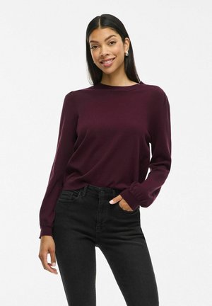 Pull maroon à manches longues avec un col rond, texture lisse et poignets côtelés, associé à un jean noir taille haute.