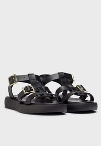 Sandalias de cuero negro con tiras entrelazadas, hebillas doradas y una suela plana de goma con una superficie texturizada.