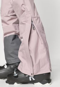 Világos rózsaszín hó nadrág szürke részletekkel. Cipzáras oldalsó nyílással, tartós anyagból, laza szabással, amely fekete síbakancsok fölött viselhető.