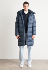 Marineblaue Daunenjacke mit Kapuze, ausgestattet mit einem durchgehenden Reißverschluss, gestepptem Design und großen Abschnitten. Getragen über einem marineblauen Pullover und hellen Jeans.