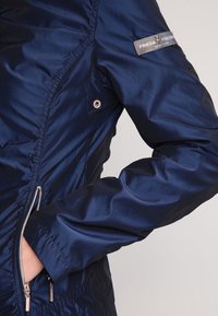 Navy blue, lightweight nylon jacket with a zipped front, side pockets, and a logo patch on the sleeve. Smooth texture, fitted design.