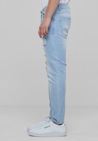 Vaaleansiniset slim fit -farkut, joissa on kulutettuja kohtia ja risareunat, valmistettu denimistä, yhdistetty valkoisiin tennareihin, joissa on vihreitä yksityiskohtia.