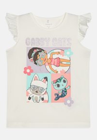 Hvit lin skjorte med volanger på ermene, prydet med tegneseriefigurer i fargerike firkanter. Teksten leser "GABBY CATS" i glitrende bokstaver.