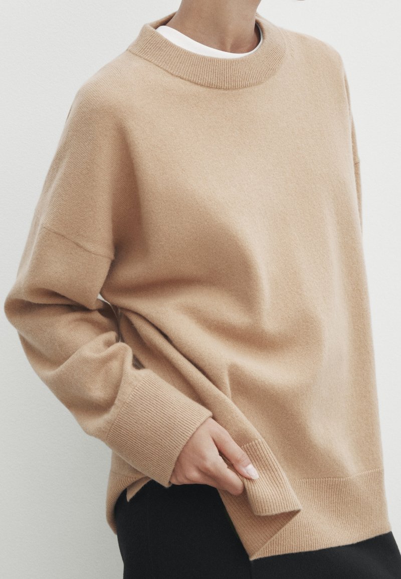 Beige kashmirtröja med hög ribbad krage, sänkta axlar och ribbade muddare, som visar upp en mjuk, texturerad yta.