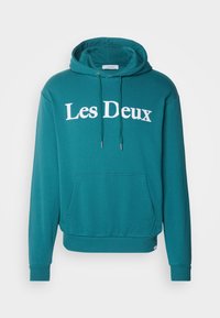 Teal hoodie with white "Les Deux" text. Features a kangaroo pocket, adjustable drawstrings, and ribbed cuffs and hem. Soft, casual fabric.