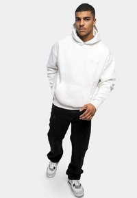 Felpa bianca con cappuccio e tasca frontale, abbinata a pantaloni neri e sneakers bianche. La texture del tessuto sembra morbida e rilassata.