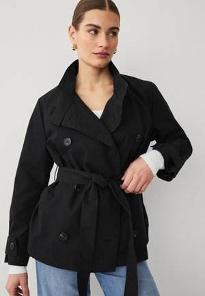 Vrouw draagt een zwart dubbelrijig jasje met riem, een wit shirt eronder, blauwe spijkerbroek en gouden oorringen, met haar in een staart.