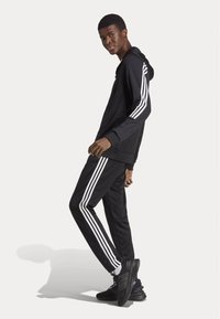 Hoodie noir et pantalon de jogging avec des rayures blanches sur les côtés, tissu texturé, associé à des chaussures de sport noires, se tenant contre un fond blanc.