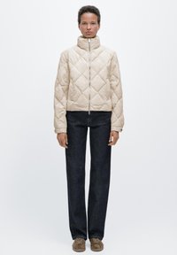 Beżowa pikowana kurtka z wysokim kołnierzem i zamkiem z przodu, zestawiona z ciemnoniebieskimi prostymi dżinsami i jasnobrązowymi trampkami.