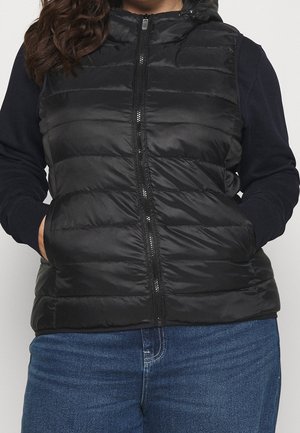 Frau trägt eine schwarze gesteppte ärmellose Kapuzenweste über einem dunklen Langarmshirt, mit den Händen in den Taschen der Weste, kombiniert mit blauen Jeans.
