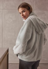 Fleece hoodie in lichtgrijs met een zachte textuur, oversized pasvorm, elastische manchetten en een capuchon met trekkoord, gezien vanaf de achterkant.