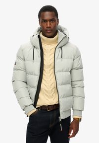 Mand iført en lys grå puffjakke over en cremefarvet kabelstrikket turtleneck sweater og mørkeblå jeans, står med den ene hånd i lommen.