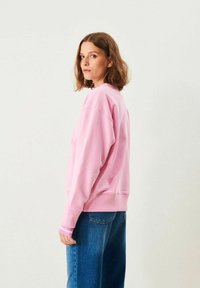 Kvinna med axellångt brunt hår, klädd i en ljusrosa sweatshirt och blå jeans, ståendes i profil mot en enfärgad bakgrund.