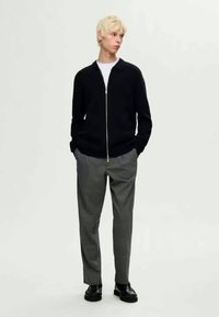 Melna apgrieztā jaka ar pieguļošu siluetu, uzvilkta virs baltas t-kreklu. Apvienota ar pelēkām ķeksētām biksēm un melniem apaviem.