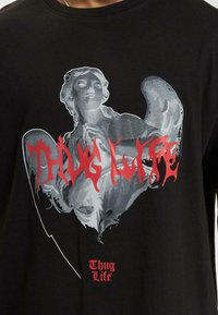 Čierne tričko s grafickým dizajnom postavy s krídlami v sivej farbe a červeným textom "THUG LIFE", vytlačeným v štýlovom písme.
