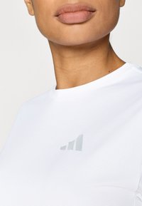 Fehér rövid ujjú sportpóló texturált anyagból, a mellkason finom szürke logóval. Kerek nyakú kialakítás.