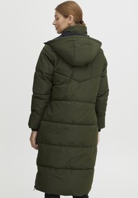 Lång vadderad kappa i mörkgrönt med huva. Har horisontella sömmar och rak skärning, tillverkad av mjukt, isolerande material.