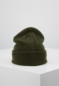 Olive green knit beanie with a folded cuff and a textured finish, featuring a small black tag with a yellow logo on the side.