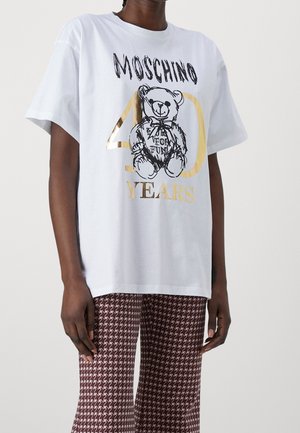 Osoba nosząca białą koszulkę Moschino z grafiką pluszowego misia i złotym tekstem "40 Years", dobraną do wzorzystych brązowo-białych spodni.