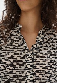 Blusa a maniche corte beige con motivo astratto nero. Presenta un colletto con bottoni e una texture del tessuto liscia.