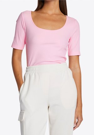 Kobieta ubrana w różowy prążkowany top z krótkim rękawem i dekoltem w serek, wsunięty w białe spodnie z wysokim stanem, elastycznym paskiem w talii oraz klapką kieszeni bocznej.