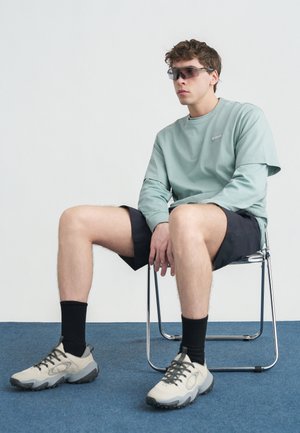 Νεαρός άνδρας με γυαλιά ηλίου, πράσινο μπλουζάκι με μακριά μανίκια, μαύρο σορτς, μαύρες κάλτσες και μπεζ αθλητικά παπούτσια, που κάθεται σε μεταλλική καρέκλα πάνω σε μπλε χαλί.