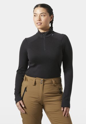 Žena v černém dlouhém zipovém topu a hnědých kalhotách, která se mírně dívá doprava, s vlasy v copy, na jednobarevném pozadí.