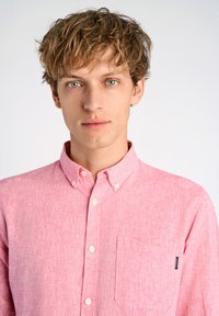 Lichtroze overhemddas in een gestructureerde stof, met een borstzak en een klein logotag aan de zijkant. Strak passend kraag.
