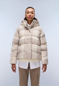 Beige rutigt pufferjacka med huva, med en texturerad ytterlager och ribbade ärmslut, bärs över en vit skjorta och beiga byxor.