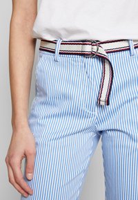 Helle blau-weiß gestreifte Hose aus Stoff, mit einem Stoffgürtel in Rot und Marineblau sowie einer silbernen, runden Schnalle.