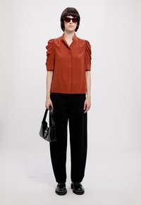 Blusa curta de cor ferrugem com mangas bufantes, combinada com calças largas pretas e sapatos pretos robustos; os acessórios incluem uma pequena bolsa preta.