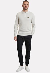Polo blanco de manga larga con cuello con cremallera y logo amarillo, combinado con pantalones cargo negros y zapatillas blancas. Textura suave, ajuste casual.