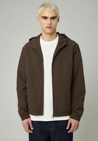 Veste brune zippée avec une capuche, des poignets élastiques et une coupe décontractée. Portée par-dessus un t-shirt blanc et un jean foncé ; texture lisse.