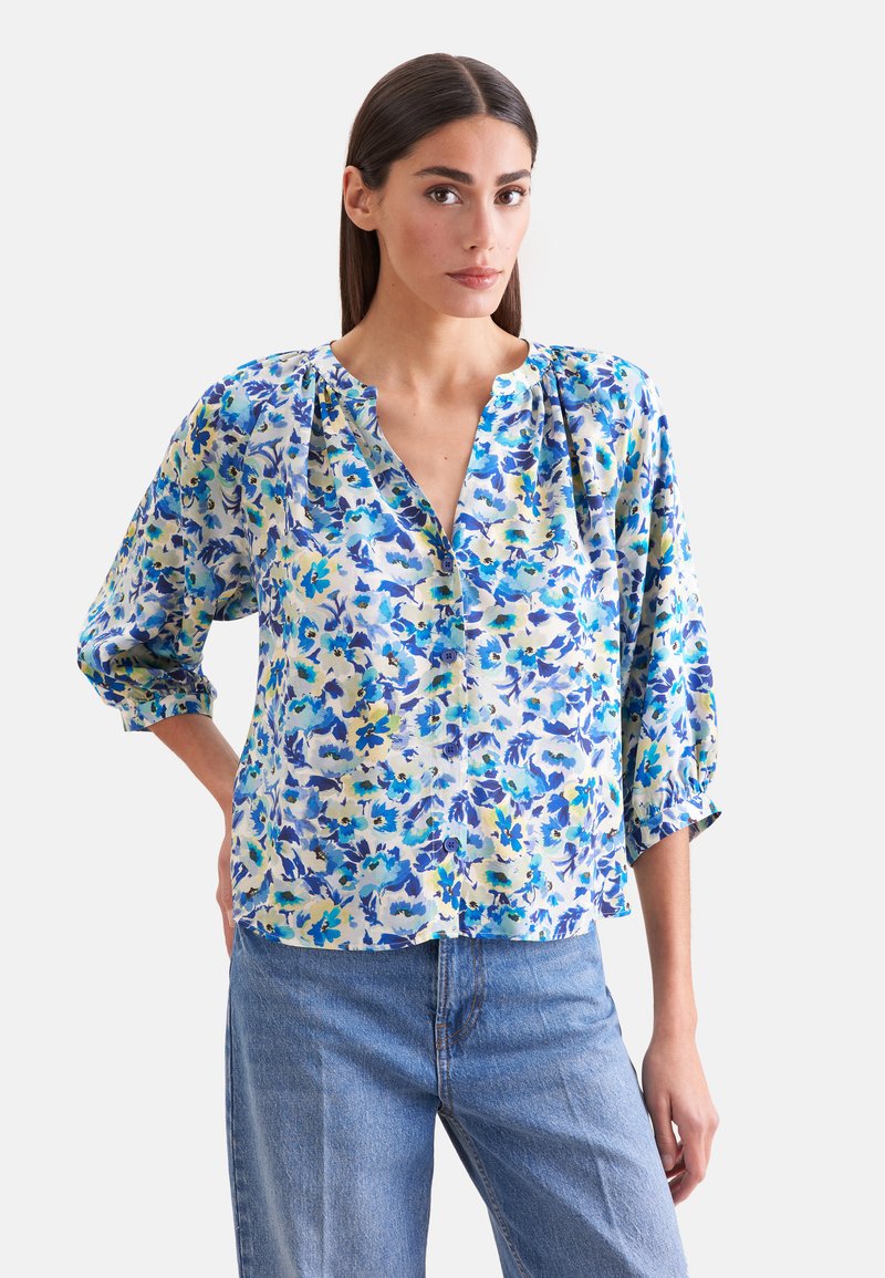 Femme aux longs cheveux foncés portant un chemisier ample à fleurs boutonné aux couleurs bleu, jaune et blanc, associé à un jean bleu clair.