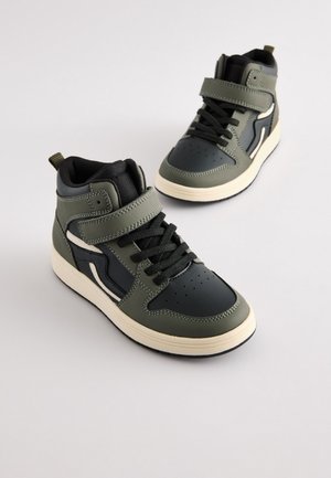 Augsti brīvā laika apavi olīvu zaļā un melnā ādā ar Velcro siksnu, melnām šņorēm, perforētu purngalu un krēmkrāsas zolīti.