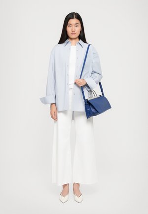 Camicia oversize azzurro chiaro, top bianco aderente, pantaloni bianchi larghi, scarpe crema, tiene una borsa blu con dettagli argentati. Tessuti con texture lisce.