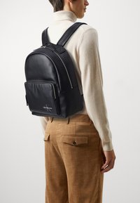 Zaino in pelle nera con forma arrotondata, tasca frontale con zip e spallacci. Presenta un logo sul davanti e una texture liscia.